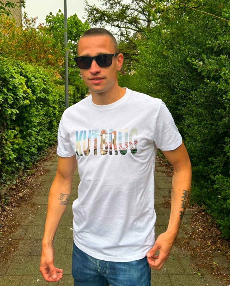 KUTBRUG, de besteller van KledingKracht op een T-shirt Jonge man in een wit t-shirt met de tekst "KUTBRUG", staand op een pad tussen bomen.