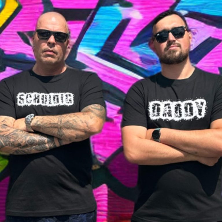 Voor onze Kledingkracht webshop, druk hier Twee mannen in zwarte T-shirts met de teksten "Schuldig" en "Daddy" staan voor een kleurrijke graffiti-muur.