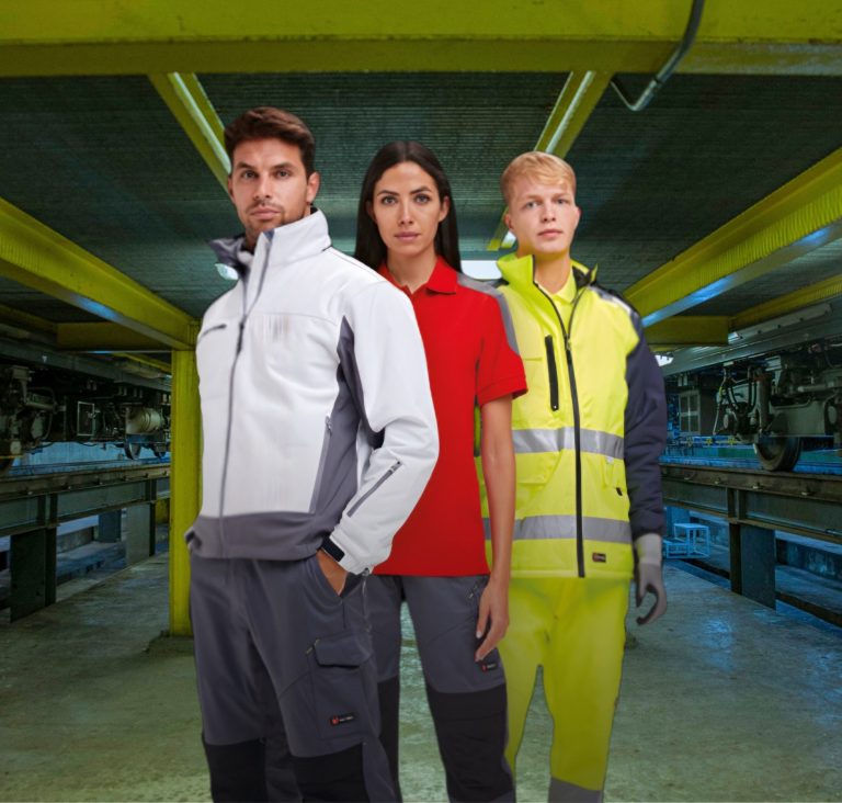 Voor bedrijfskleding, druk hier. Drie mensen in werkoutfits staan in een industriële omgeving met gele verlichting.